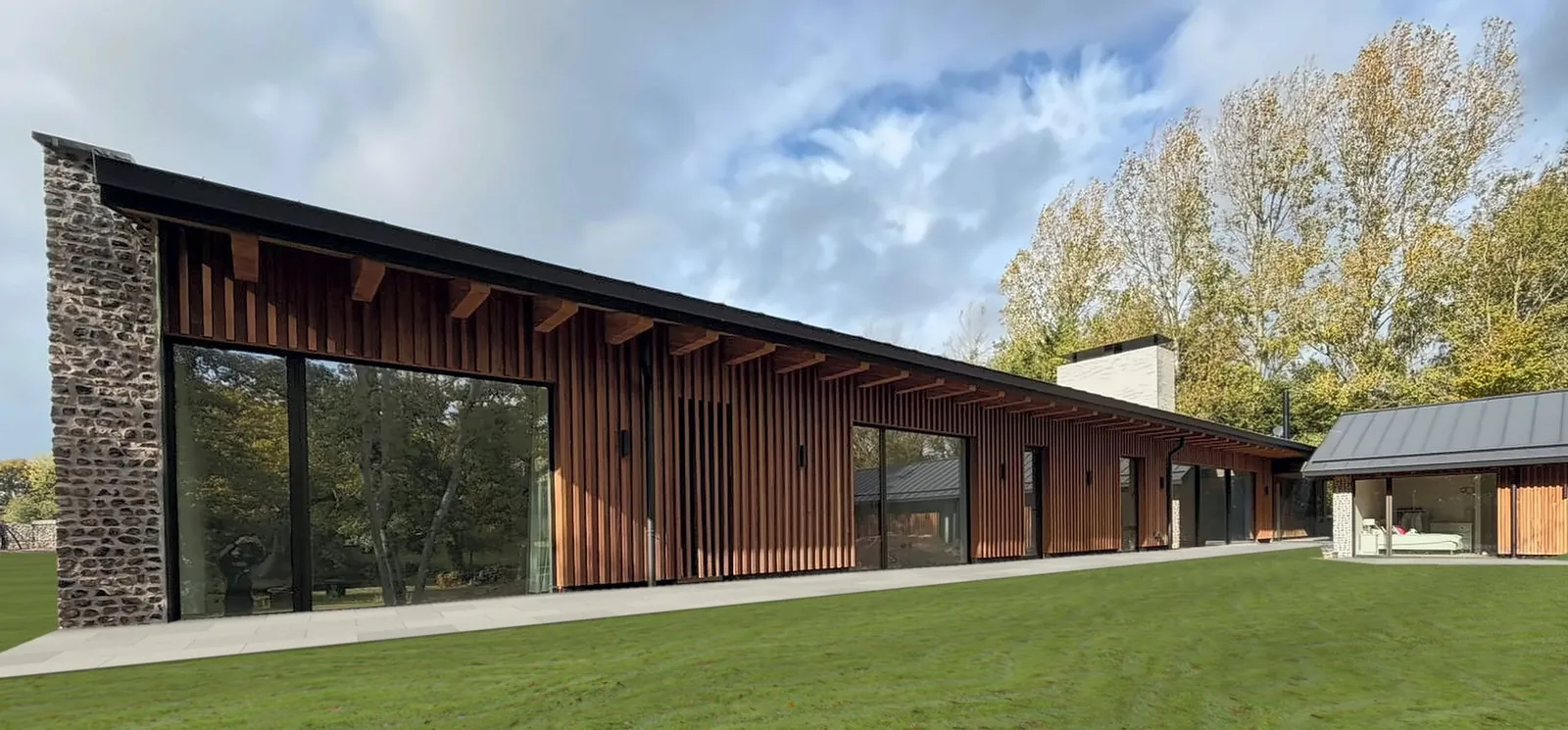 Long, single-storey house with flint walls, a zinc roof and full-height glazing, set on a lawn at the edge of woodland